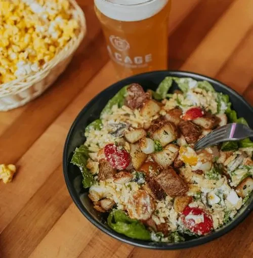 Black bowl of hearty salad, beer, and popcorn on wooden table.