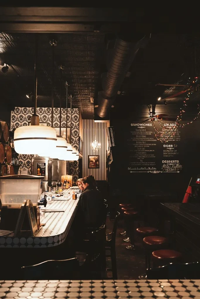 Dimly lit bar interior with white-tiled counter, pendant lights, and chalkboard menu.