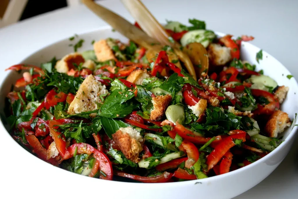 Fresh salad with peppers, cucumber, croutons, and herbs in a white bowl.