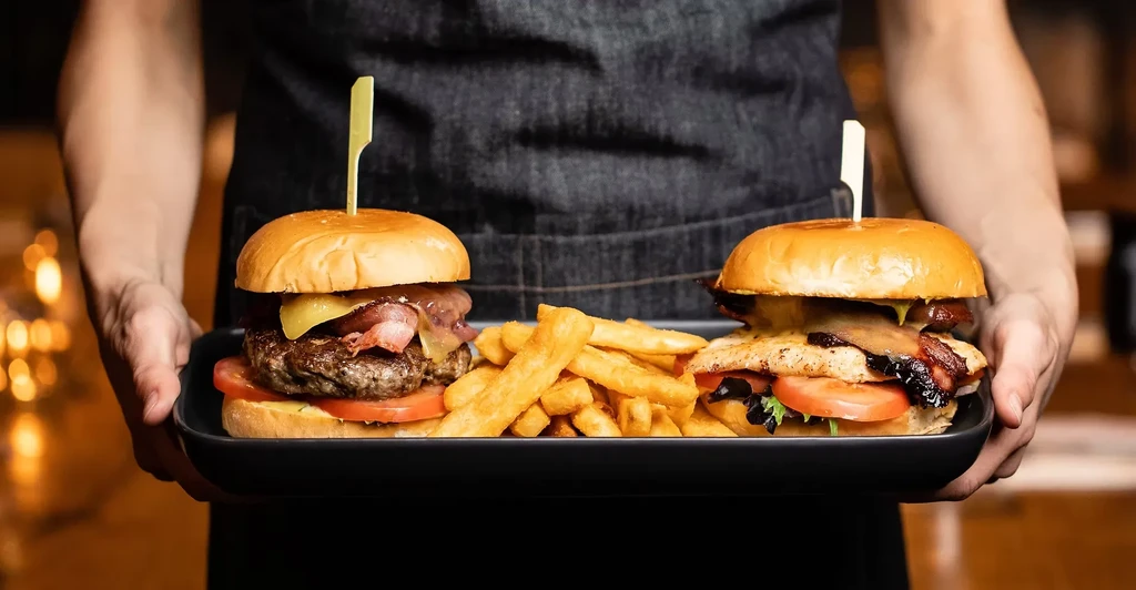 Close-up: Hands holding two gourmet burgers and fries on a black tray.
