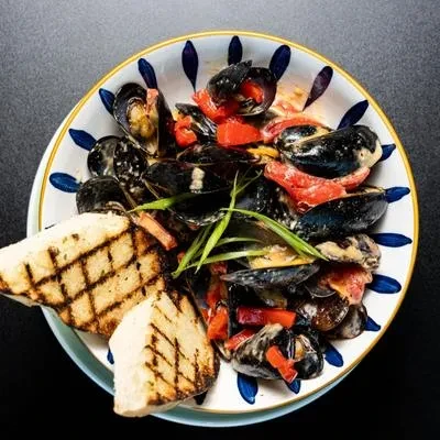 Top-down view of cooked mussels, red vegetables, green herbs, grilled bread.