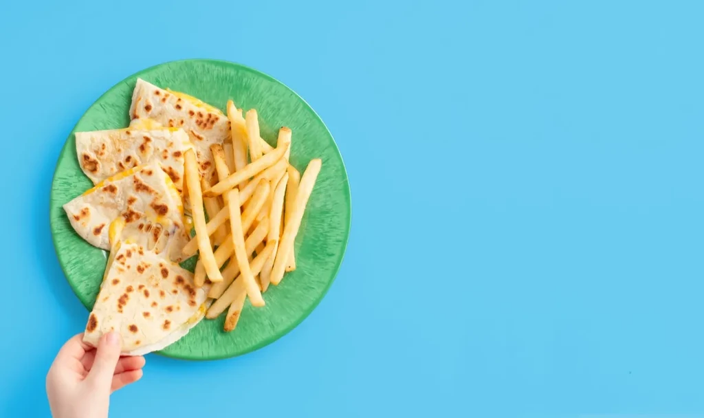Hand taking cheesy quesadilla and fries from green plate, blue background.