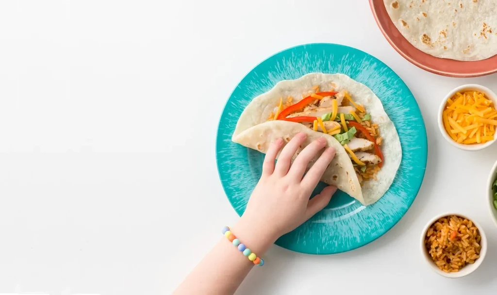 Overhead: child's hand folds a chicken taco with rice, cheese, tortillas.
