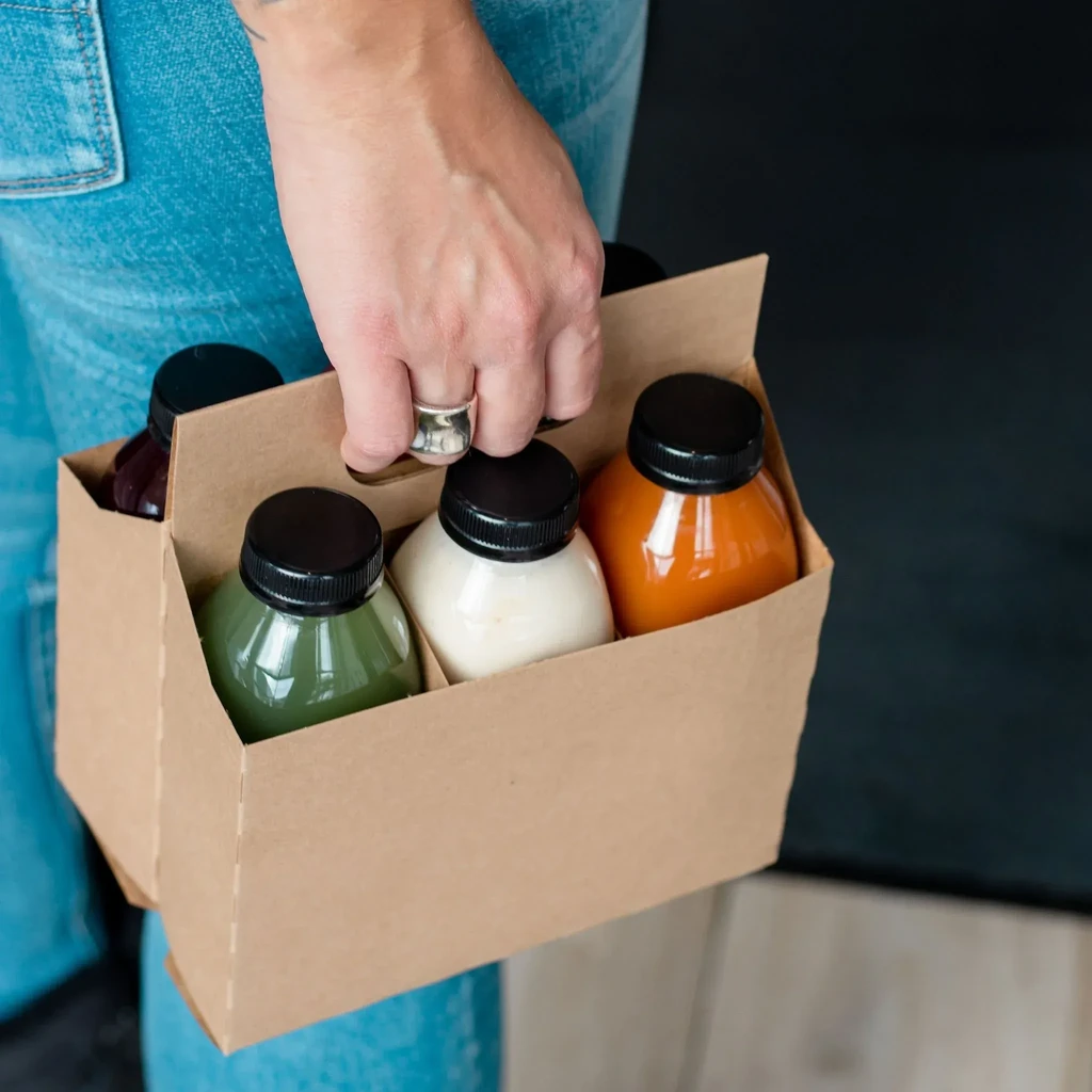Hand holding a brown cardboard carrier with six colorful bottled drinks.