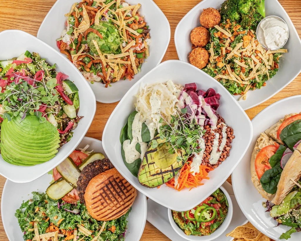 Overhead view of diverse healthy salads, grain bowls, burger on wooden table.
