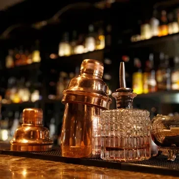 Copper shakers, glass decanter on a dark bar with blurred liquor bottles.