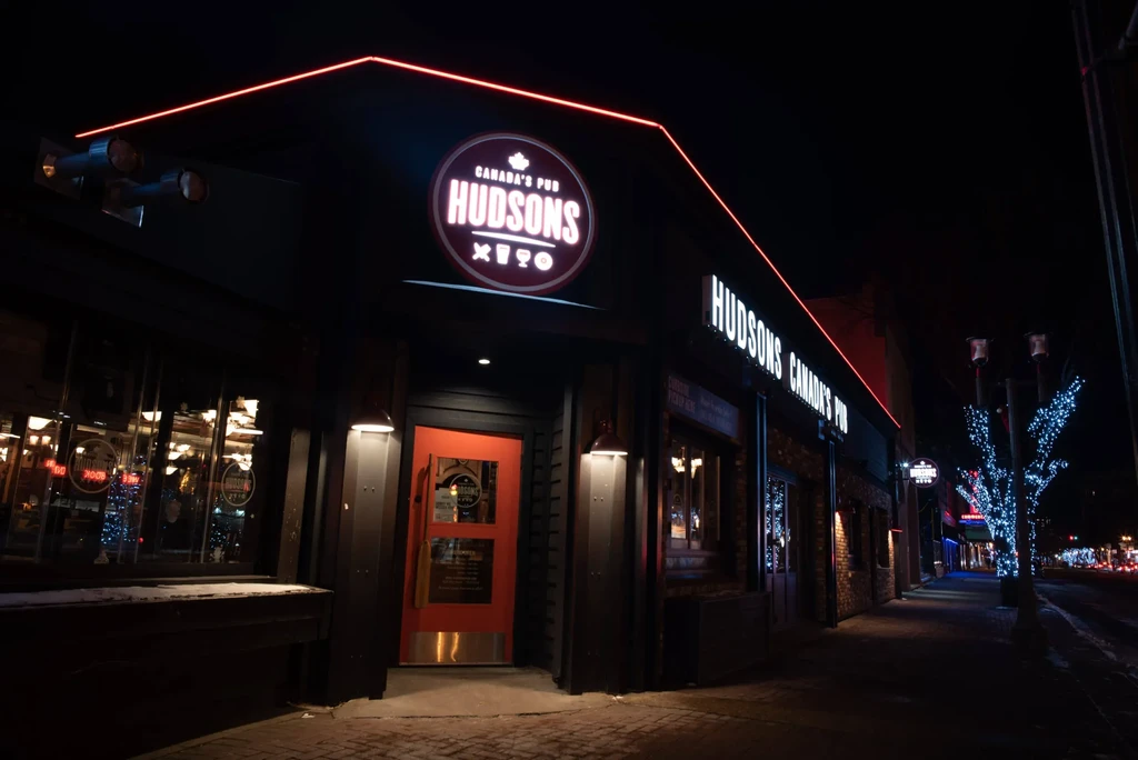 Hudsons Canada's Pub at night, illuminated by bright neon and tree lights.