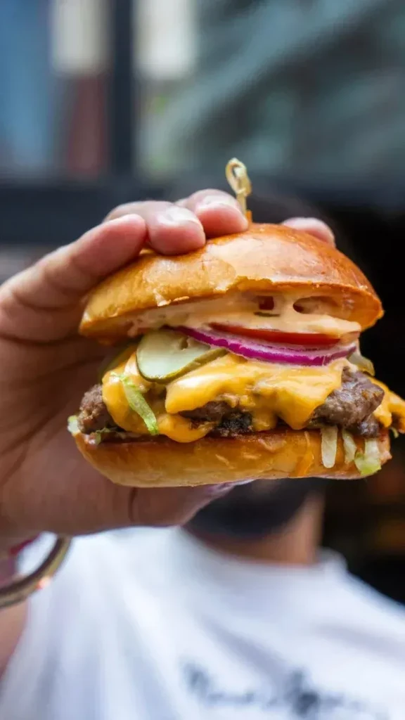 Hand holding a large double-patty burger with cheese, lettuce, onion, and pickles.