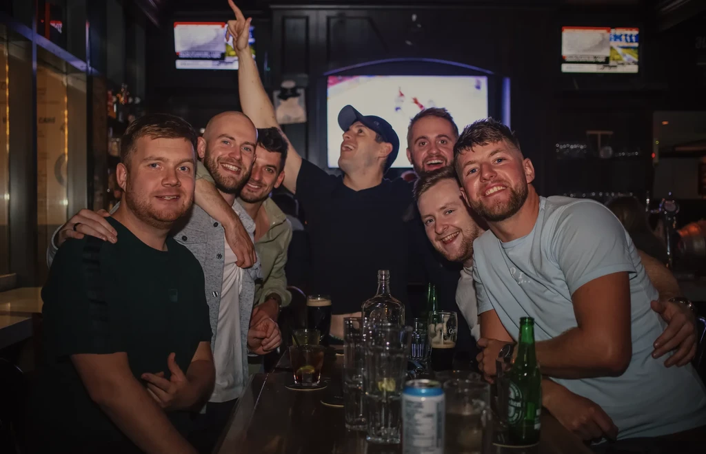 Seven men celebrating together around a bar with drinks.