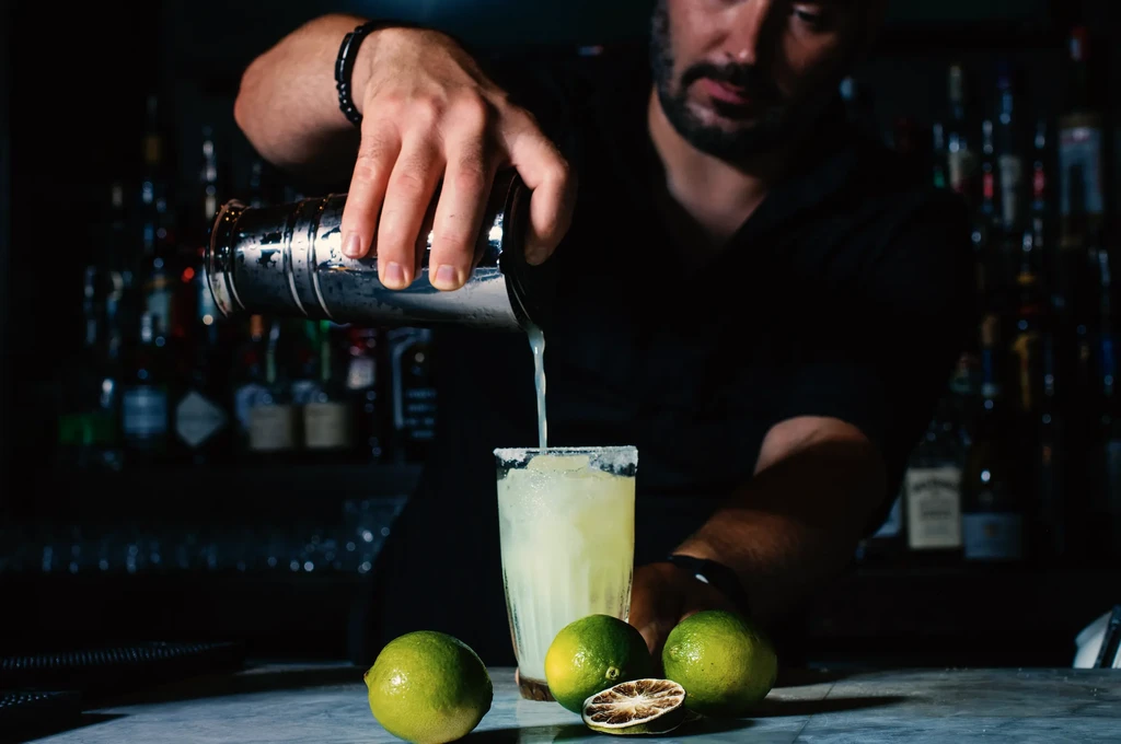 Bartender pours light drink from shaker into salt-rimmed glass with limes.