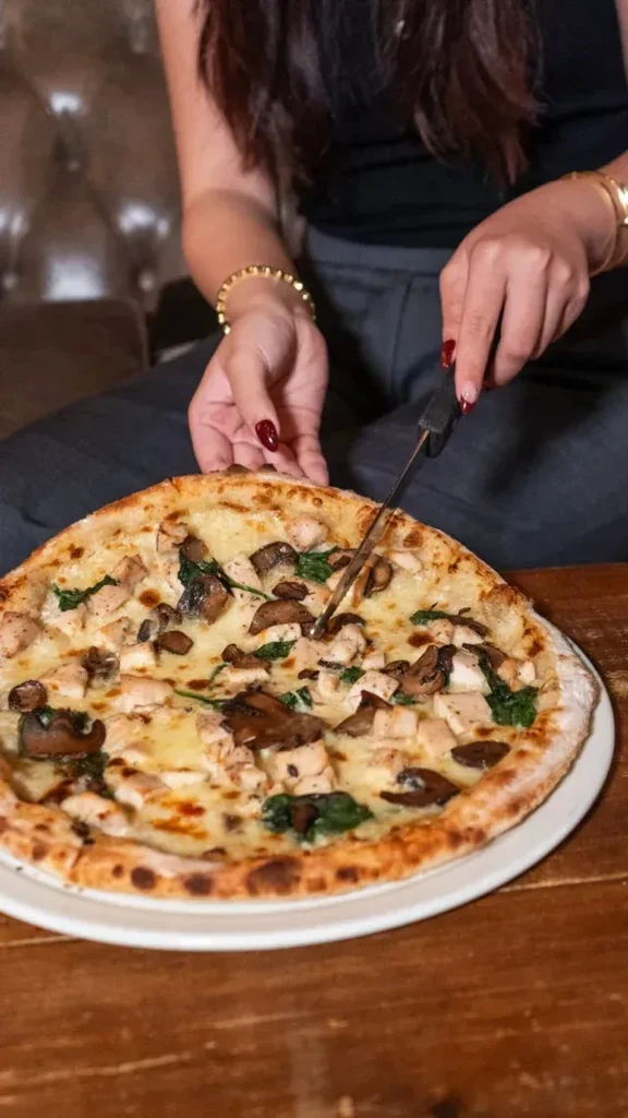 Hands cutting a white-sauce pizza with chicken, mushrooms, spinach.
