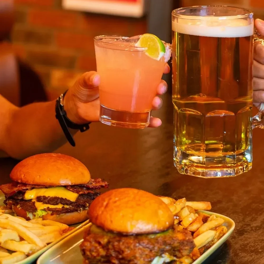 Hands toasting a cocktail and beer over a cheeseburger and chicken sandwich.