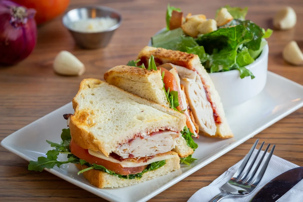 Club sandwich halves, green salad, fork, and knife on a wooden table.