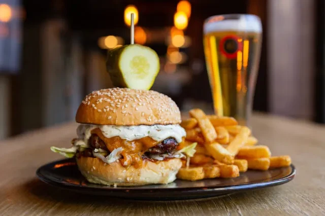 Well-lit gourmet burger, golden fries, pickle skewer, and blurred beer.