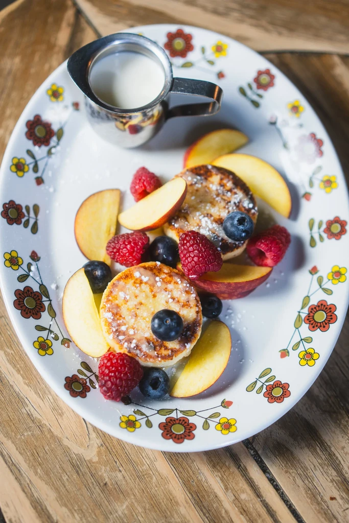 Golden pancakes, berries, and apple slices on a floral plate with creamer.