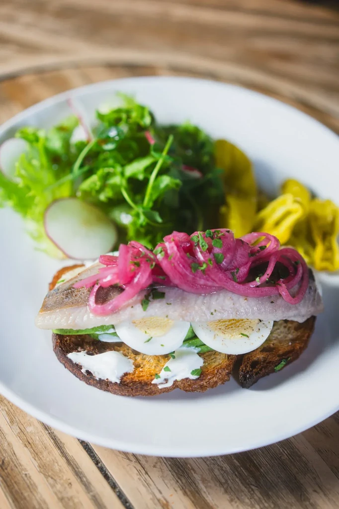 Plated open-faced sandwich with fish, egg, pickled onions, and greens.