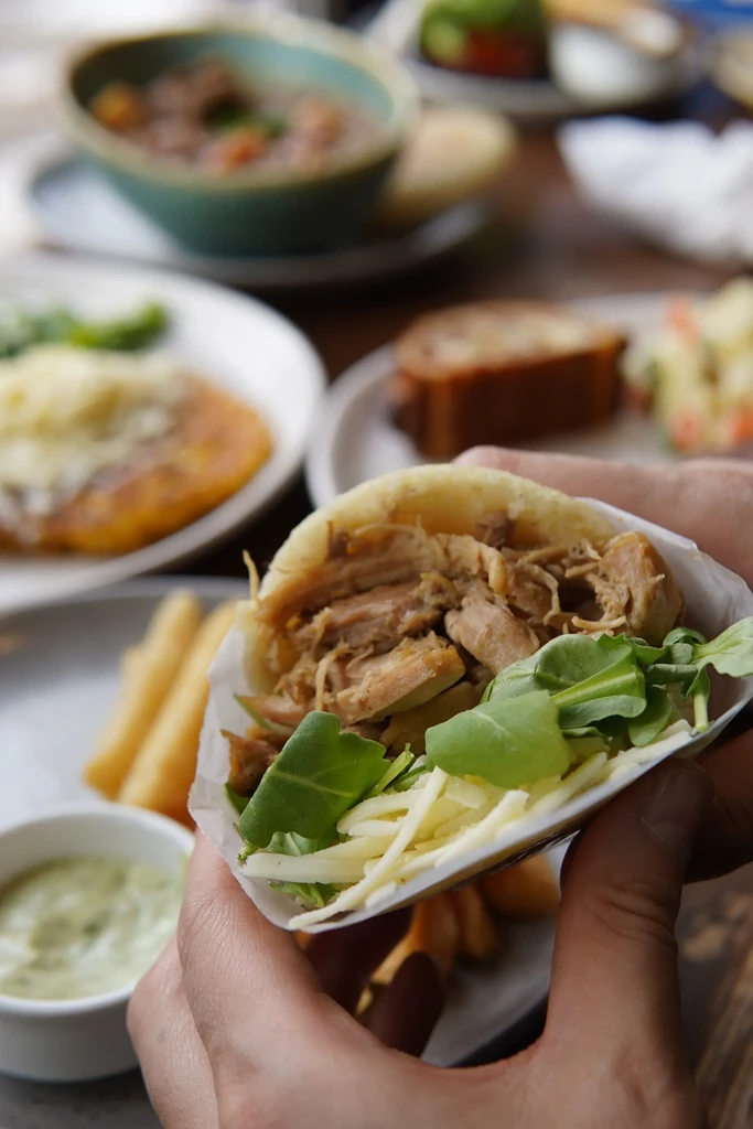 Hands hold open flatbread revealing shredded meat, cheese, and leafy greens.