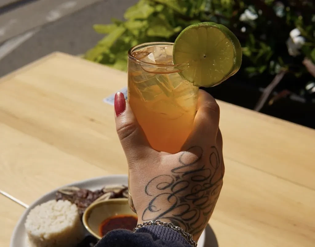 Hand with script tattoo holds iced amber drink with lime.