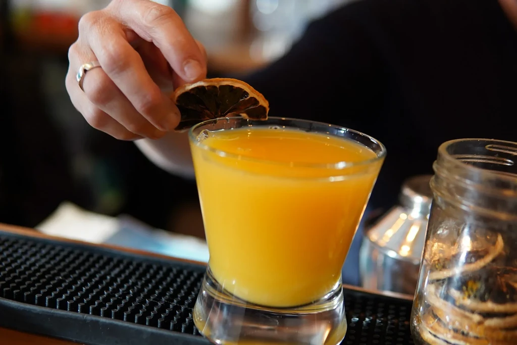 Hand garnishes vibrant orange drink with a dried citrus slice.