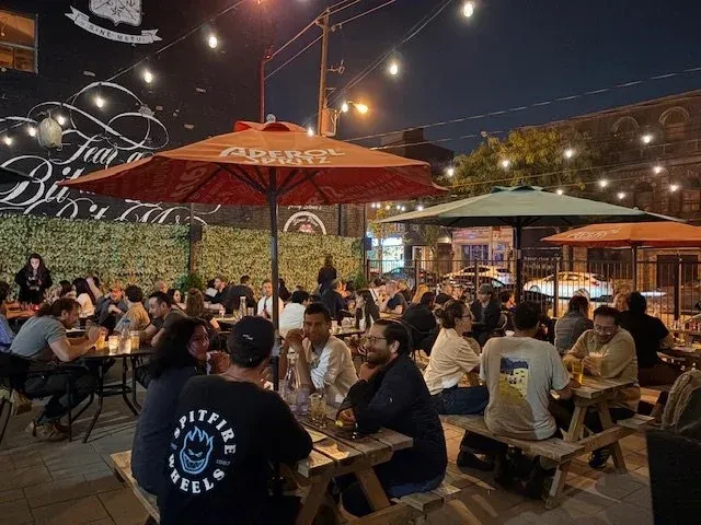 Nighttime urban patio, patrons under string lights, Aperol umbrella visible.