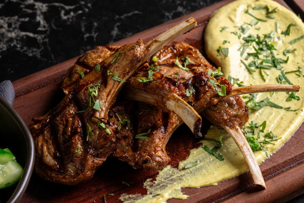Close-up of glossy grilled lamb chops with herbs, creamy sauce, sliced cucumbers.
