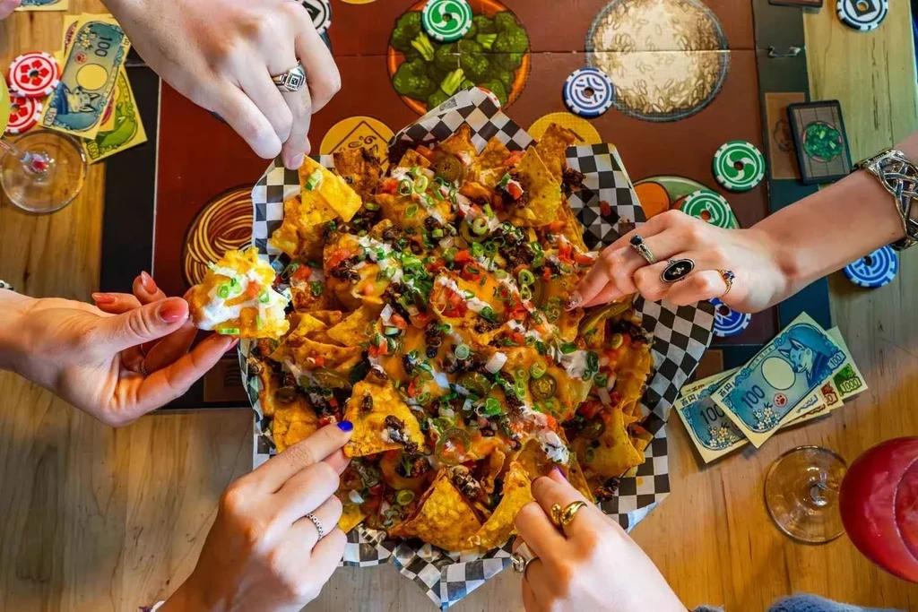 Hands reach for loaded nachos and drinks around a board game.
