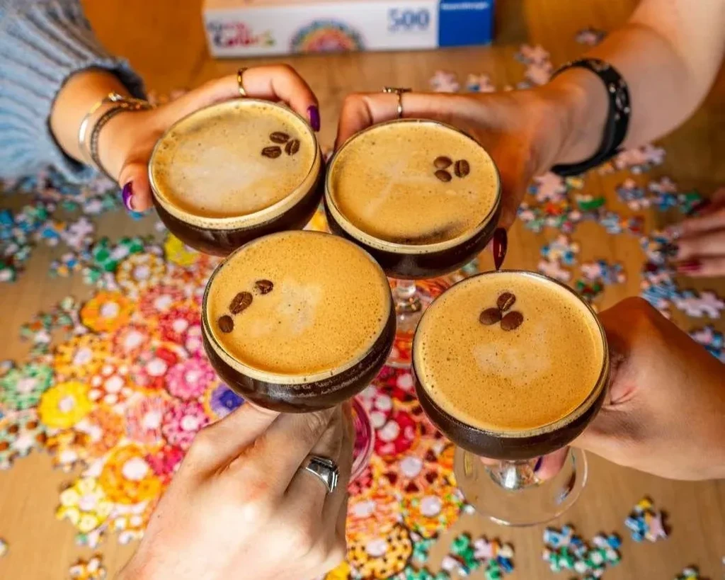 Four hands toast with Espresso Martinis over a colorful scattered puzzle.