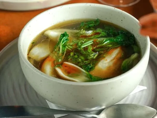 Clear broth soup with dumplings and greens in a white bowl.