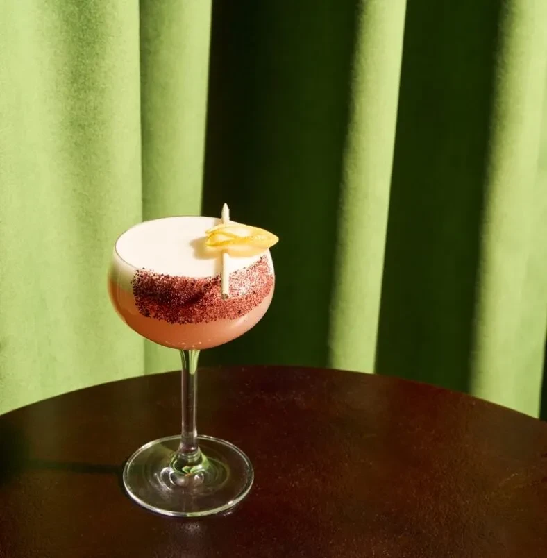 Pinkish-orange cocktail, white foam, spice-dusted rim, citrus curl.