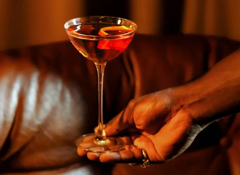 Reddish-brown cocktail, orange peel in coupe glass, held by ringed hand.