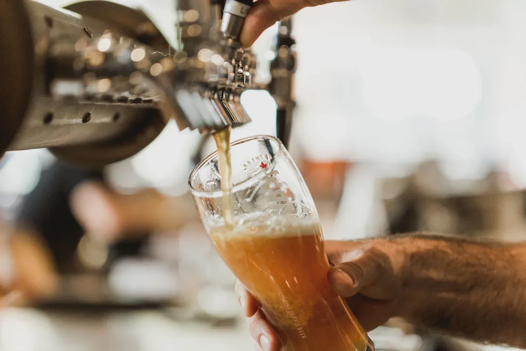 Hands pour foamy golden beer from a tap into a branded glass.