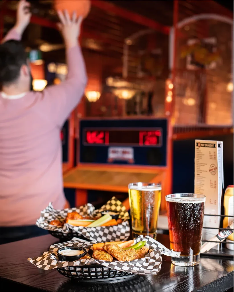 Two baskets of fried food, two beers; person plays arcade basketball.