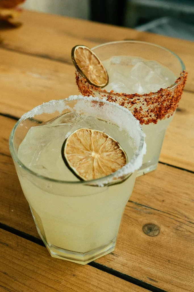 Two iced drinks with lime, salt, and spice on a wood table.