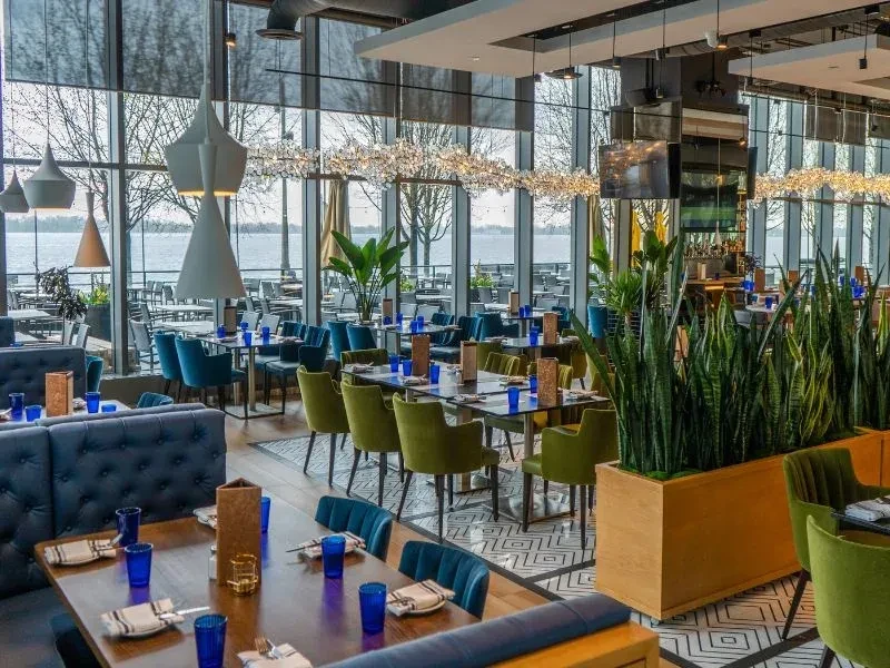 Modern restaurant interior with waterfront view, plush seating, and plants.