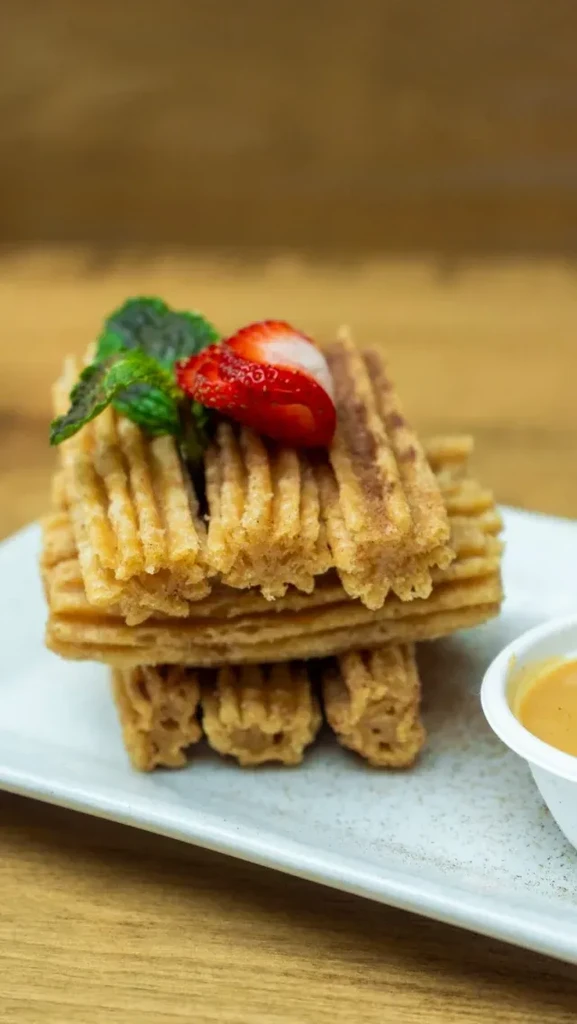 Close-up of golden-brown churros stacked with strawberry, mint, and dipping sauce.