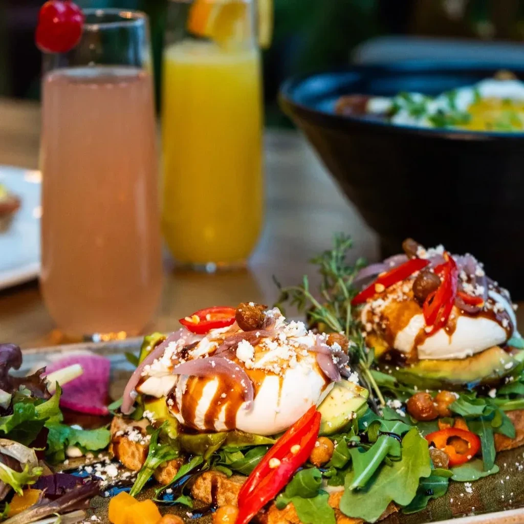 Close-up of avocado toast with poached eggs, cheese, and two drinks.