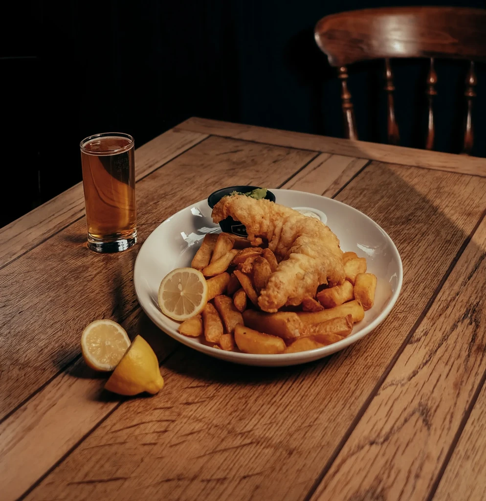 Fish and chips on wooden table with lemon, green sauce, and drink.