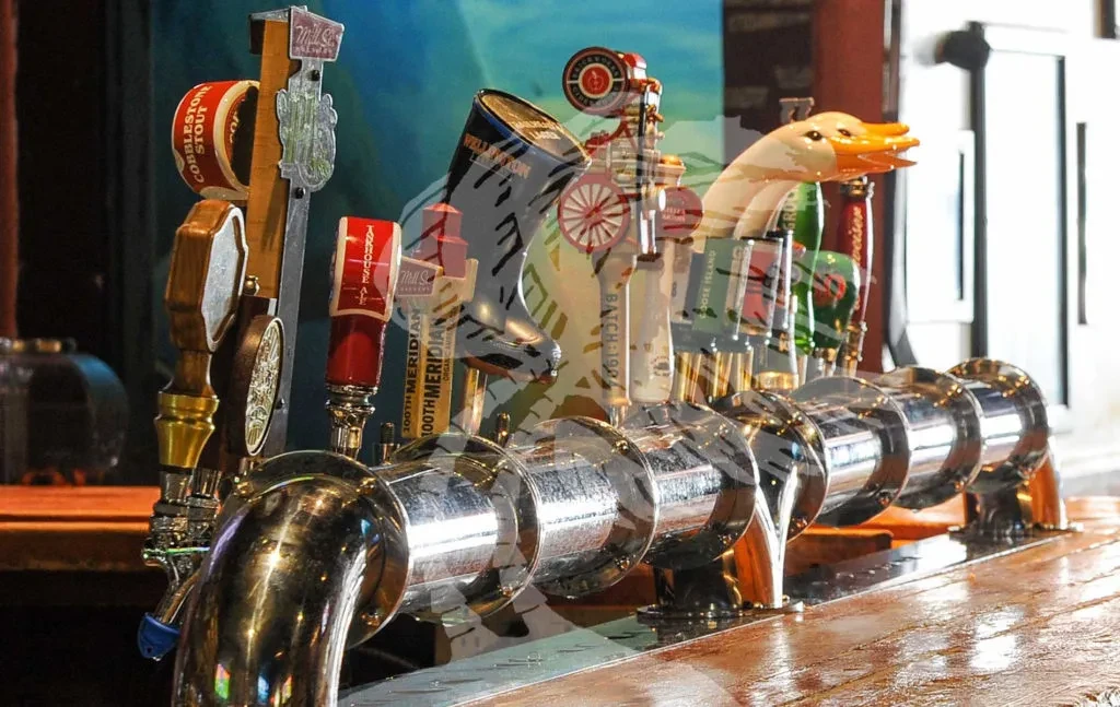Polished chrome beer taps with diverse handles line a wooden bar.