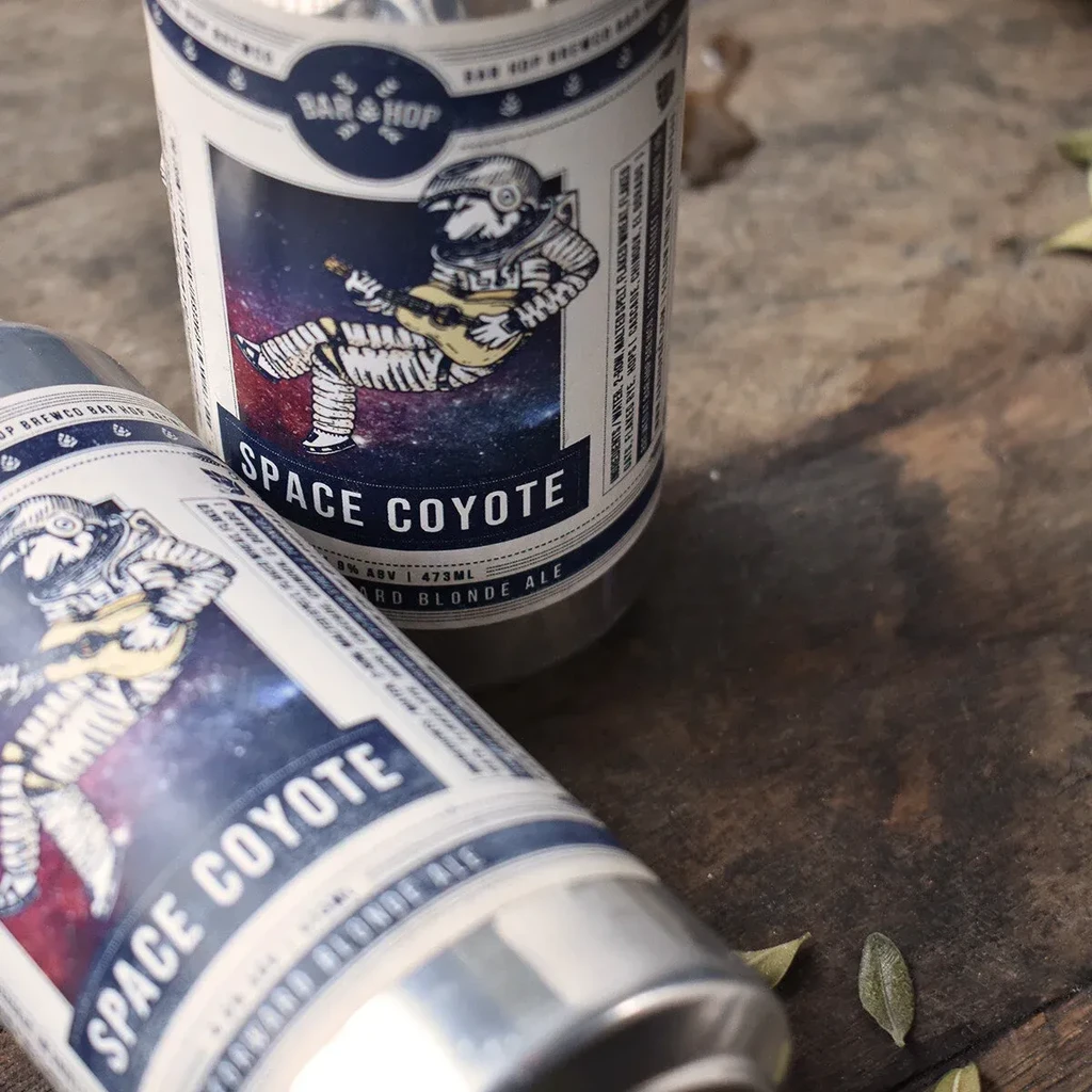 Two silver "SPACE COYOTE" beer cans with astronaut labels on wood.