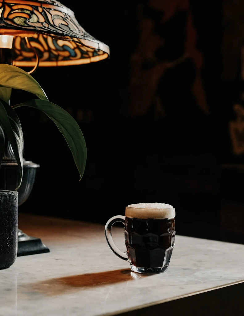 Frothy dark drink in faceted mug on marble, near Tiffany lamp.