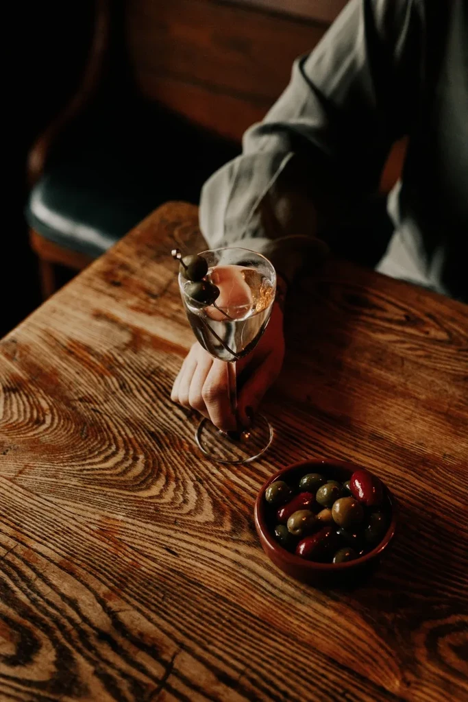 Hand holds olive martini, bowl of olives on rustic table.