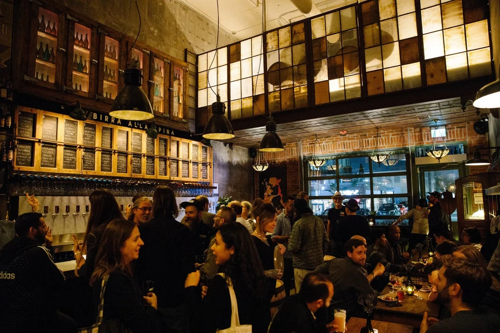 Bustling bar interior with patrons socializing around beer taps.