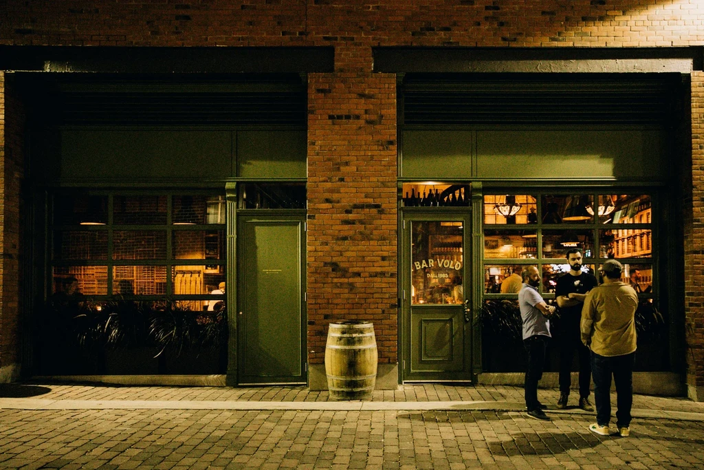Night view of BAR VOLO brick building with warm lights and three men.