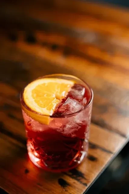 Dark red cocktail with ice and an orange slice on a wooden bar.