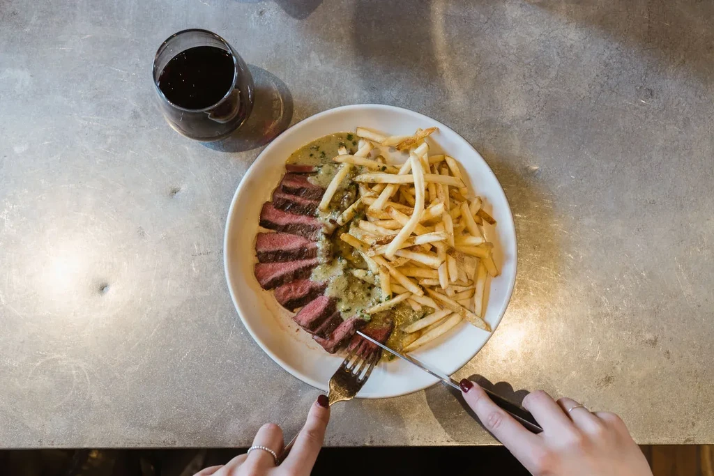THOUGHTS:
The user wants an alt-text between 7 and 12 words.
It needs to be factual, concise, and descriptive, with no marketing language.
I need to focus on the key elements: steak, fries, wine, and someone cutting the food.

Let's break down the descript