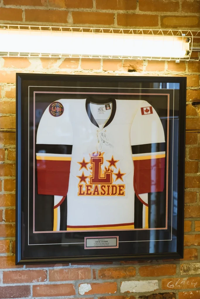 Framed white Leaside hockey jersey with 'L' on a brick wall.