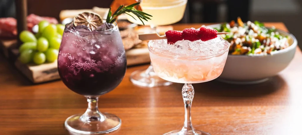 Two professionally prepared cocktails, purple and peach, on a wooden table.