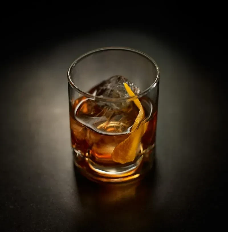 Amber cocktail with ice and twisted orange peel in a rocks glass.