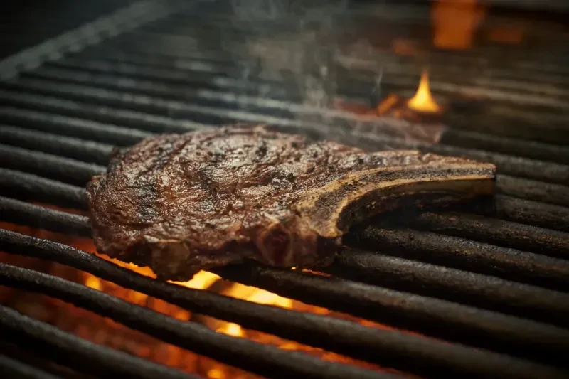 Large bone-in steak grilling over glowing embers and open flames.