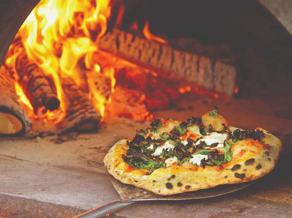 Wood-fired pizza with greens on a peel, inside a glowing oven.